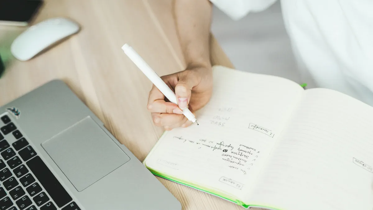 person writing notes in notebook beside a laptop